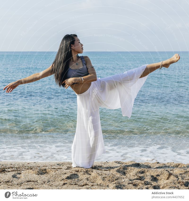 Asiatische Frau mit angehobenem Bein übt Yoga am Strand Bein angehoben Gleichgewicht üben Gesunder Lebensstil Dehnung Wellness Vitalität Meeresufer asiatisch