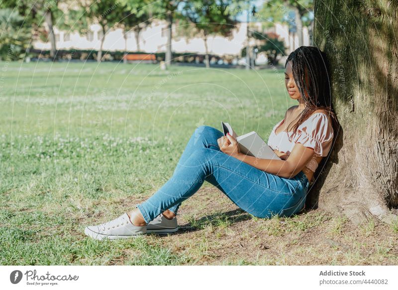 Nachdenkliche ethnische Frau mit Buch, die sich im Park ausruht nachdenken wehmütig reflektierend trendy tausendjährig Rasen Schnüffler Top Lehrbuch Literatur