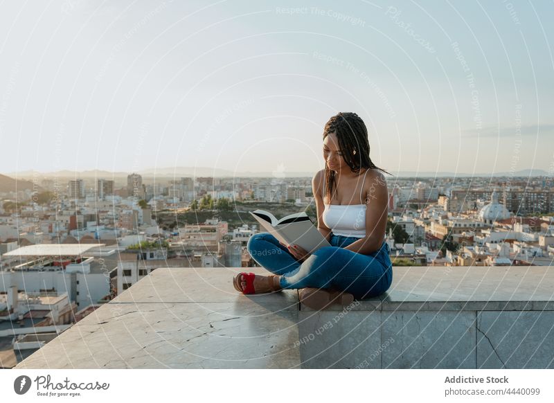 Coole schwarze Frau, die auf einem Dach in der Stadt ein Buch liest lesen Lehrbuch freie Zeit Literatur trendy Beine gekreuzt achtsam Dachterrasse Großstadt