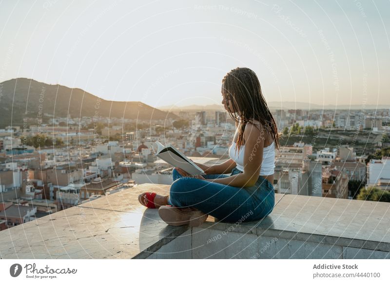 Coole schwarze Frau, die auf einem Dach in der Stadt ein Buch liest lesen Lehrbuch freie Zeit Literatur trendy Beine gekreuzt achtsam Dachterrasse Großstadt