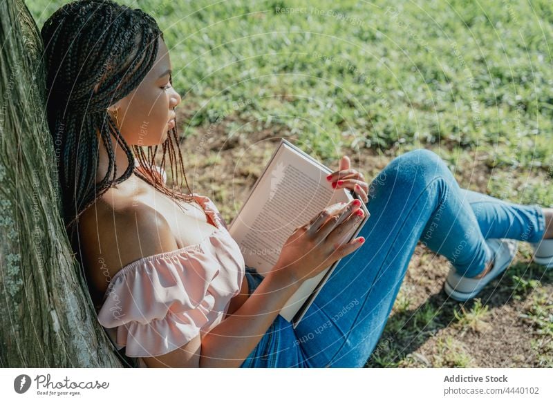 Nachdenkliche ethnische Frau mit Buch, die sich im Park ausruht nachdenken wehmütig reflektierend trendy tausendjährig Rasen Schnüffler Top Lehrbuch Literatur