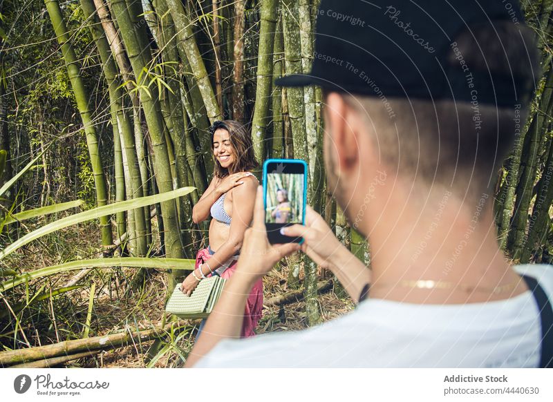 Crop-Tourist, der seine fröhliche Freundin mit dem Smartphone fotografiert Frau fotografieren heiter Haare berühren Tourismus Bambus benutzend Apparatur Lächeln