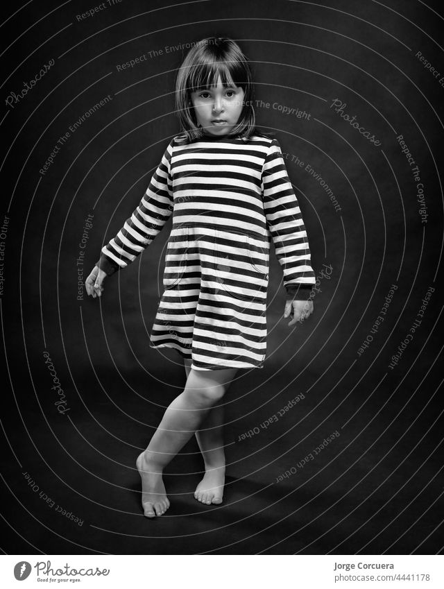 4 Jahre altes Mädchen posiert elegant in einem schwarz-weiß gestreiften Kleid. Monochromer fester schwarzer Hintergrund. 4-5 Jahre alternative Pose Baby-Mädchen