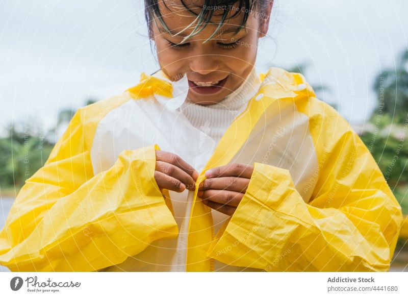 Lächelndes asiatisches Mädchen knöpft sich einen Regenmantel im Park zu zuknöpfen Kindheit freundlich charmant angenehm regnerisch Wetter heiter Inhalt achtsam