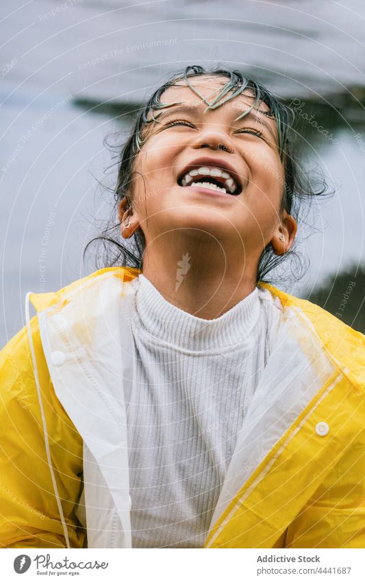 Fröhliches ethnisches Mädchen im Regenmantel hat Spaß im Park Spaß haben heiter spielen Kindheit sorgenfrei Porträt freie Zeit Himmel asiatisch froh Glück