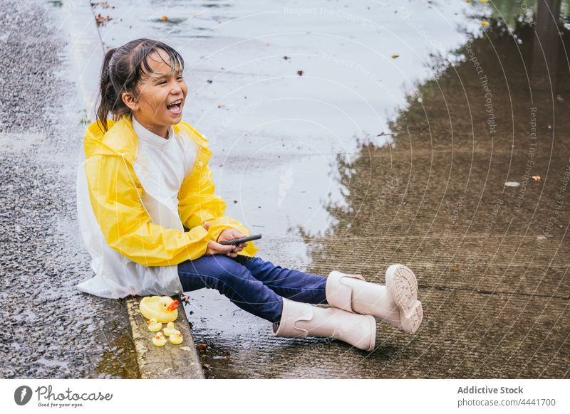Glückliches asiatisches Mädchen mit Smartphone auf dem Bürgersteig bei Regenwetter Lachen sorgenfrei Kindheit Spielzeug Pfütze ruhen Straßenbelag regnerisch