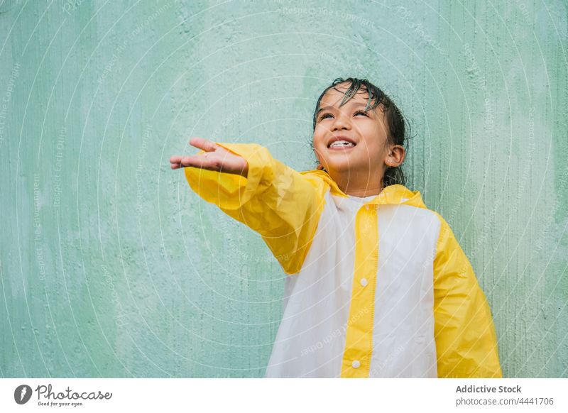 Lächelnde asiatische Mädchen fangen regen Tropfen gegen raue Wand Regen mit der Hand nach oben heiter Kindheit genießen Regenmantel herzlich charmant ethnisch