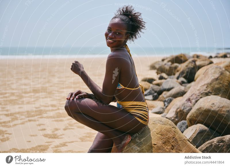 Lächelnde schwarze Frau in Badekleidung auf einem Felsbrocken an der Küste ruhend Badebekleidung Körper feminin sanft Afro-Look natürlich Meeresküste heiter