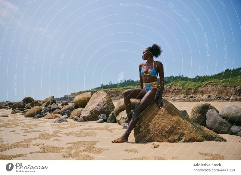 Lächelnde schwarze Frau in Badekleidung auf einem Felsbrocken an der Küste ruhend Badebekleidung Körper feminin sanft Afro-Look natürlich Meeresküste heiter