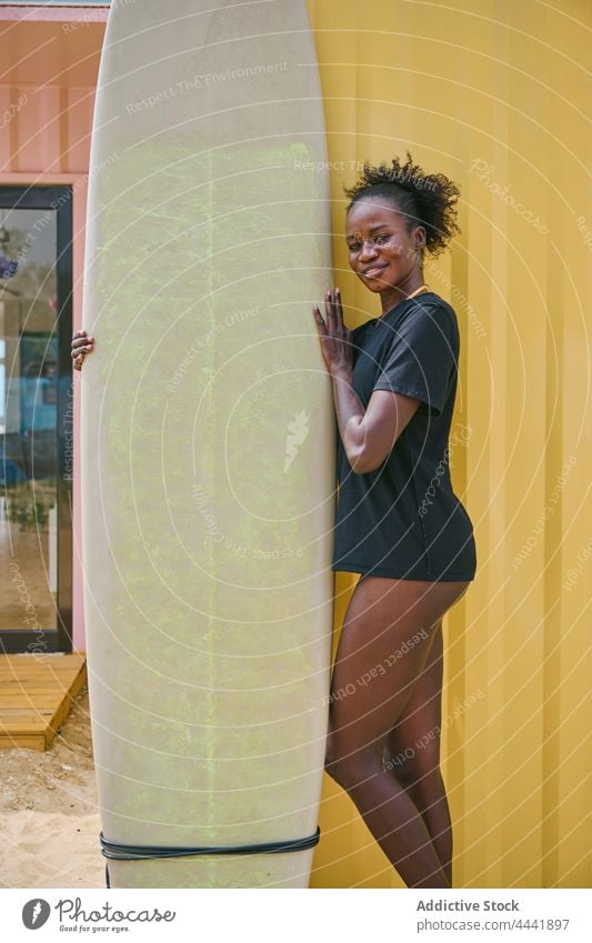 Lächelnder schwarzer Surfer mit Longboard am Ufer Sport Surfen nachdenken Körper Frau Küste Strand heiter Bikini T-Shirt Wand Sportlerin ethnisch Afroamerikaner