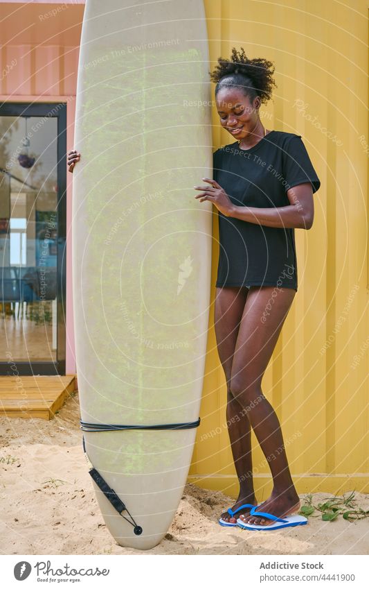 Lächelnder schwarzer Surfer mit Longboard am Ufer Sport Surfen nachdenken Körper Frau Küste Strand heiter Bikini T-Shirt Wand Sportlerin ethnisch Afroamerikaner