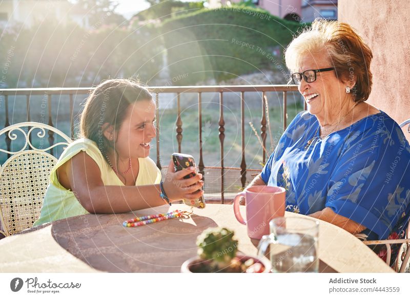 Positives Mädchen zeigt der fröhlichen Großmutter auf der Terrasse im Sonnenlicht ihr Smartphone Enkelin Glück Lächeln heiter zeigen manifestieren Frau Kind