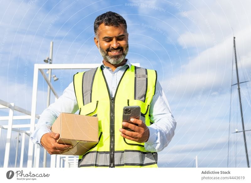 Lächelnder ethnischer Arbeiter mit Kiste, der auf seinem Smartphone surft Mann Glück benutzend führen Kollo froh Versand Uniform Freude zuschauen männlich Beruf