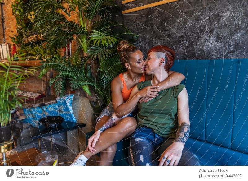 Lächelndes homosexuelles Paar mit Getränk küsst sich auf dem Sofa zu Hause Freundin Kuss Partnerschaft Liebe gleichgeschlechtlich Umarmen trinken reden Liege