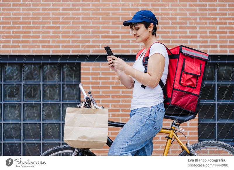 Fahrer Frau trägt roten Rucksack liefern Lebensmittel auf einem Fahrrad, Überprüfung der Bestellung mit Smartphone, während auf der Straße in der Stadt stehen. Konzept eines Lieferdienstes. Nachhaltiger Transport