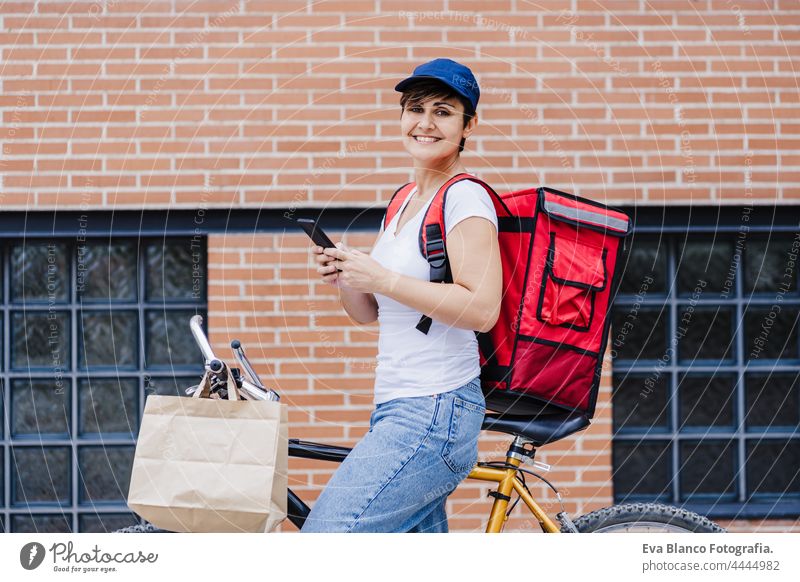 lächelnd Fahrer Frau trägt roten Rucksack liefern Lebensmittel auf einem Fahrrad, Überprüfung der Bestellung mit Smartphone, während auf der Straße in der Stadt stehen. Konzept eines Lieferdienstes. Nachhaltiger Transport