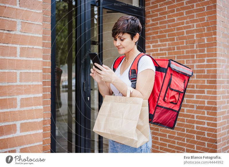 Junge Reiterin mit rotem Rucksack, die eine Pappschachtel mit Lebensmitteln in der Stadt hält. Konzept eines Lieferservices. Frau Überprüfung der Bestellung im Mobiltelefon