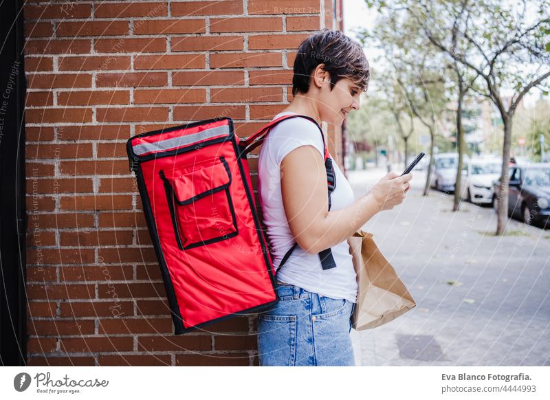 Seitenansicht des jungen Fahrers Frau trägt roten Rucksack hält Papptasche von Lebensmitteln in der Stadt. Lieferservice Konzept. Frau überprüft Bestellung im Handy