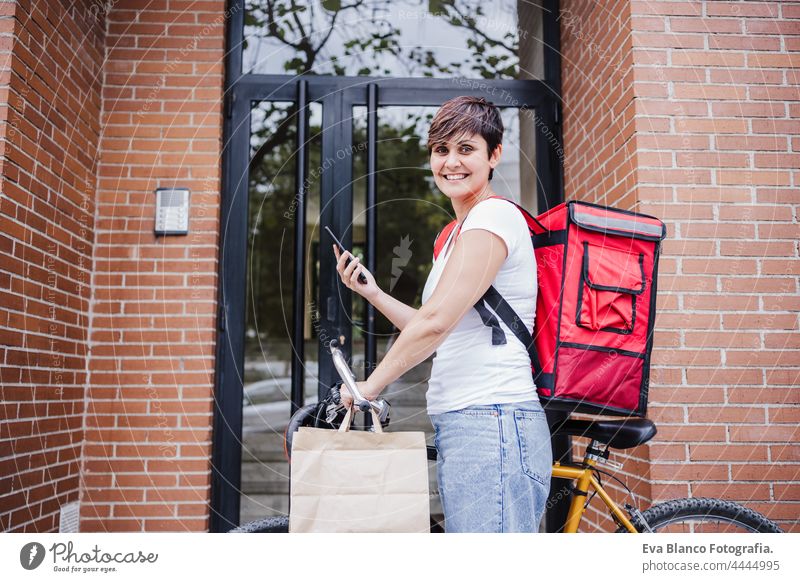 lächelnd jungen Fahrer Frau trägt roten Rucksack hält Papptasche von Lebensmitteln in der Stadt. Lieferservice-Konzept. Frau Überprüfung der Bestellung in Mobiltelefon