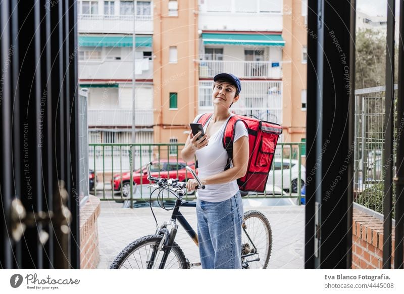 Junge Fahrerin mit rotem Rucksack, die Essen auf einem Fahrrad ausliefert und die Bestellung mit einem Smartphone überprüft, während sie am Straßentor in der Stadt steht. Konzept eines Lieferdienstes. Nachhaltiger Transport