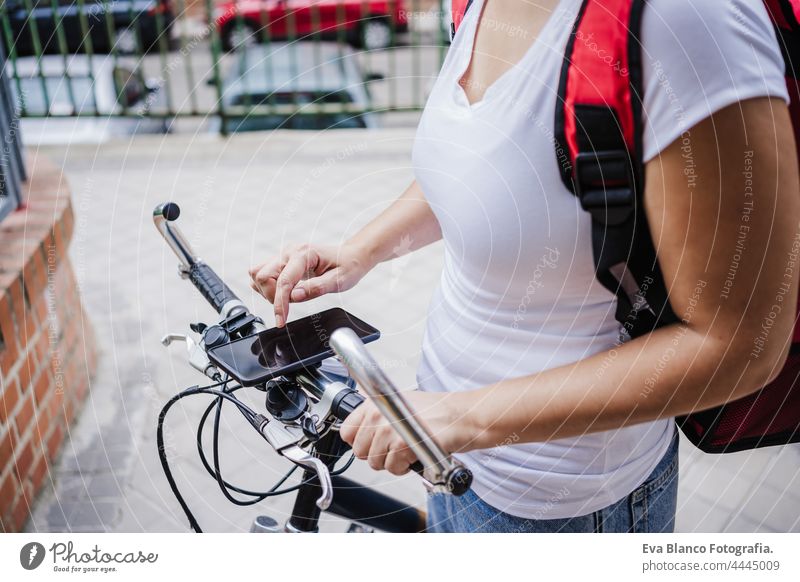 Nahaufnahme von Young Rider Frau trägt roten Rucksack liefern Lebensmittel auf einem Fahrrad, Überprüfung der Bestellung mit Smartphone, während auf der Straße Tor in der Stadt stehen. Konzept eines Lieferdienstes. Nachhaltiger Transport