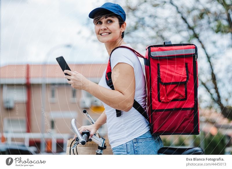 glücklicher Fahrer Frau trägt roten Rucksack liefern Lebensmittel auf einem Fahrrad, Überprüfung der Bestellung mit Smartphone, während auf der Straße in der Stadt stehen. Konzept eines Lieferdienstes. Nachhaltiger Transport