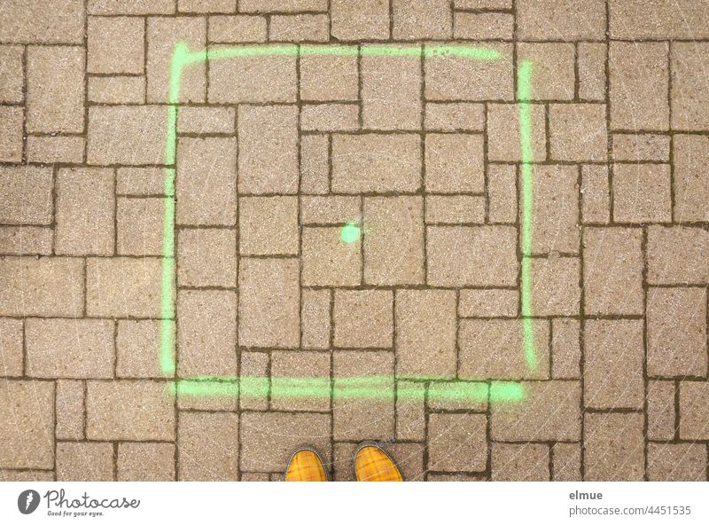 ein mit grüner Farbe aufgespraytes Quadrat und ein Punkt auf einer gepflasterten Fläche und gelbe Schuhspitzen in der Draufsicht / Platzhalter Pflaster