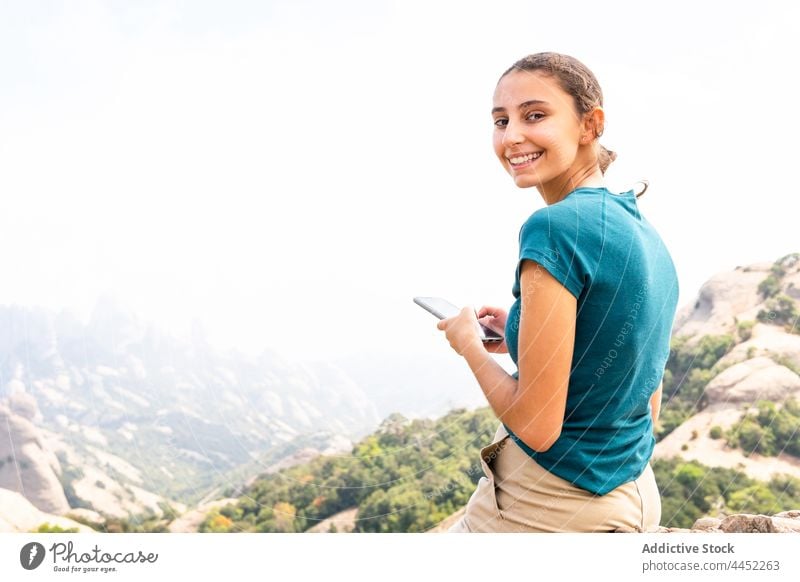 Lächelnde Frau, die während eines Ausflugs in den Bergen ihr Smartphone benutzt Tourist benutzend Nachricht Berge u. Gebirge Hochland erkunden positiv felsig