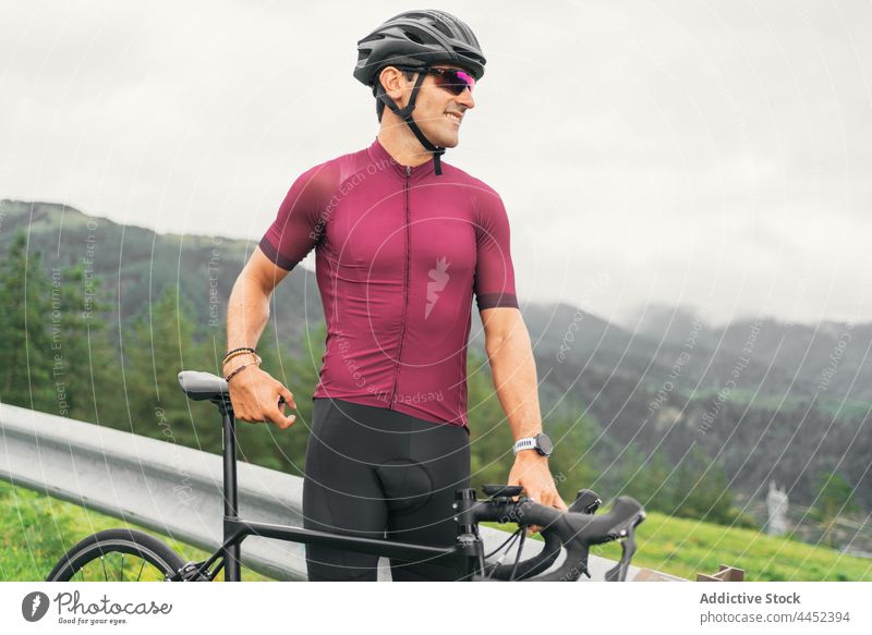 Lächelnder Radfahrer mit Schutzhelm auf dem Fahrrad auf der Fahrbahn Sport Fahrradfahren Sonnenbrille Mann Landschaft Porträt kreativ Design Straße behüten