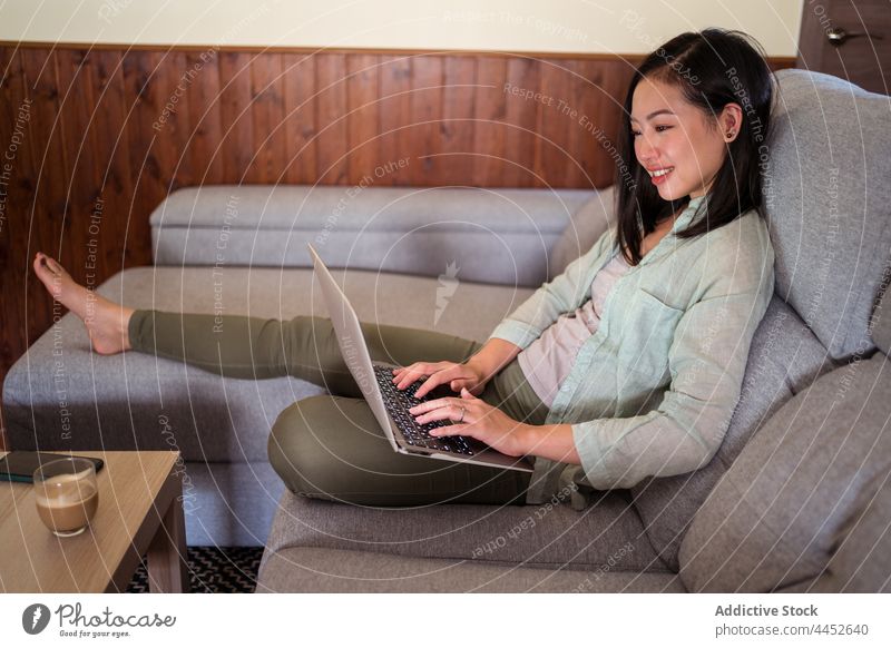Asiatischer Freiberufler tippt auf Laptop auf Couch gegen Latte Tippen Arbeit Internet Projekt Lächeln Sofa Frau benutzend Glück Apparatur interessiert Gerät