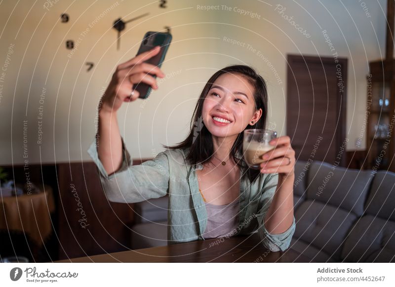 Lächelnde asiatische Frau, die ein Selfie mit Milchkaffee auf dem Smartphone macht Latte Laptop Kaffee Sofa Haus benutzend Apparatur freie Zeit heiter Inhalt