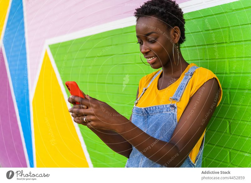 Lächelnde schwarze Frau, die in der Nähe einer bunten Wand eine SMS auf ihrem Smartphone schreibt benutzend zuschauen hell Surfen Stil positiv Textnachricht