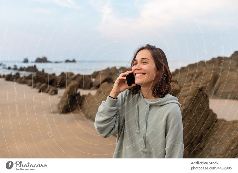 Fröhliche Frau, die an einer felsigen Küste mit ihrem Smartphone spricht reden heiter Meeresküste Felsen Natur freie Zeit wolkig benutzend Apparatur Himmel
