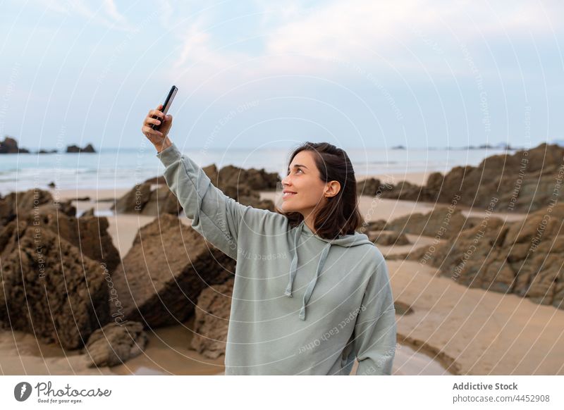 Lächelnde Frau, die ein Selfie mit ihrem Smartphone am felsigen Strand macht Selbstportrait Meeresküste Felsen Gedächtnis Moment Himmel Freizeit benutzend