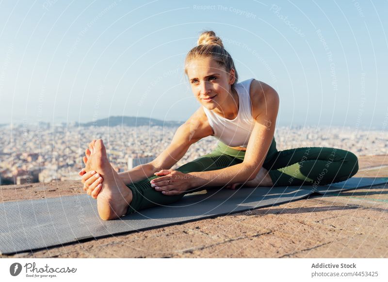 Frau streckt Bein auf Yogamatte in der Stadt Vorwärtsbeuge Dehnung Gesunder Lebensstil Tastfuß Wellness Vitalität Dachterrasse Blauer Himmel Großstadt Pose üben