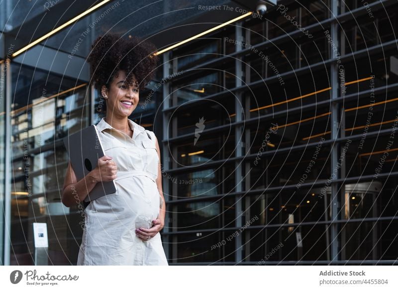 Lächelnde schwarze Frau mit Laptop kommt aus einem Bürogebäude schwanger führen mütterlich vorwegnehmen erwarten Turm Glück Bauch Apparatur Gerät digital Freude