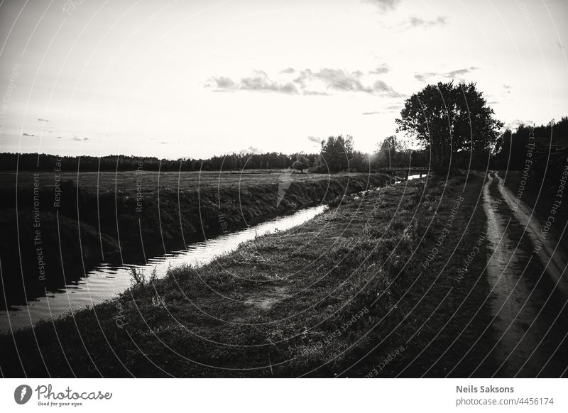 schöne schwarze und weiße abstrakte Lettland Landschaft. Dirt Sandstraße, Wasser fließt in geraden Fluss mit sauberen Banken, einige Bäume in Seite