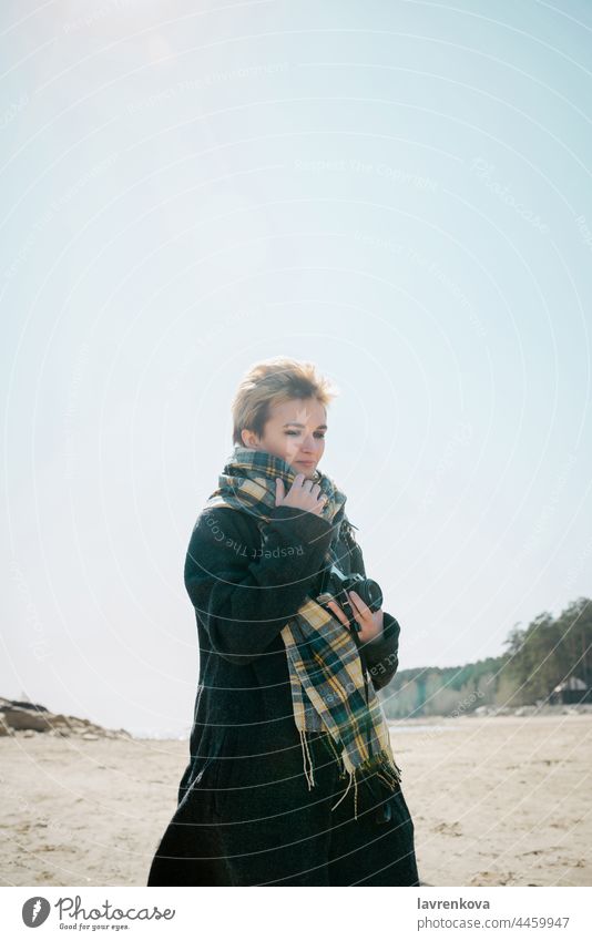 Junge Frau in Mantel und Schal mit Kamera an einem Strand bei kaltem Wetter, Erwachsener Herbst blond Kaukasier Schönwetter Brille Glück Landschaft Lifestyle