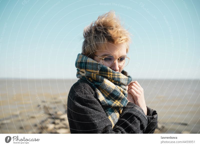 Junge Frau mit Brille wärmt sich an einem Strand allein Mantel Herbst Porträt jung Urlaub Gesicht Kaukasier Natur im Freien Seeküste Himmel reisen Wasser kalt