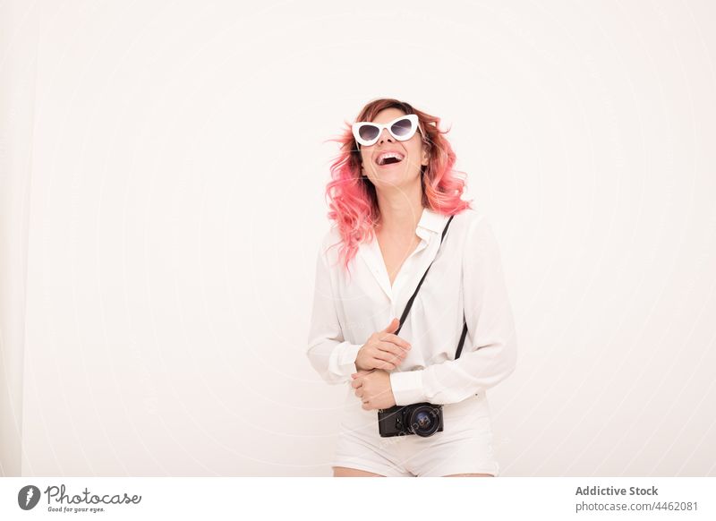Fröhliche Frau mit Sonnenbrille steht mit Fotokamera vor einer hellen Wand Stil trendy Fotoapparat Glück Lächeln Vorschein heiter positiv Outfit Raum Fotograf
