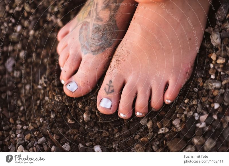 tätowierte Füße am Strand, Ankertattoo auf dem Zeh, Nagellack, nackte Füße auf Kies Tattoo shark Haifisch strand Boden Frauenfüße Damenfuß feminin Menschenfuß