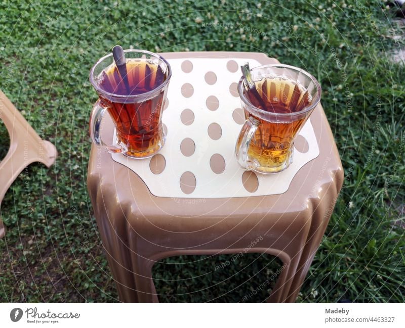 Türkische Teegläser mit Teelöffel und Tee auf einem Plastikhocker in Braun und Beige auf grünem Rasen in einem Garten im Dorf Maksudiye in der Provinz Sakarya in der Türkei