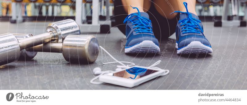 Unbekannter Sportler sitzt mit Hanteln und Handy auf dem Boden des Fitnessstudios Transparente Mobile unkenntlich Fuß Turnschuh Kopfhörer Panorama Smartphone