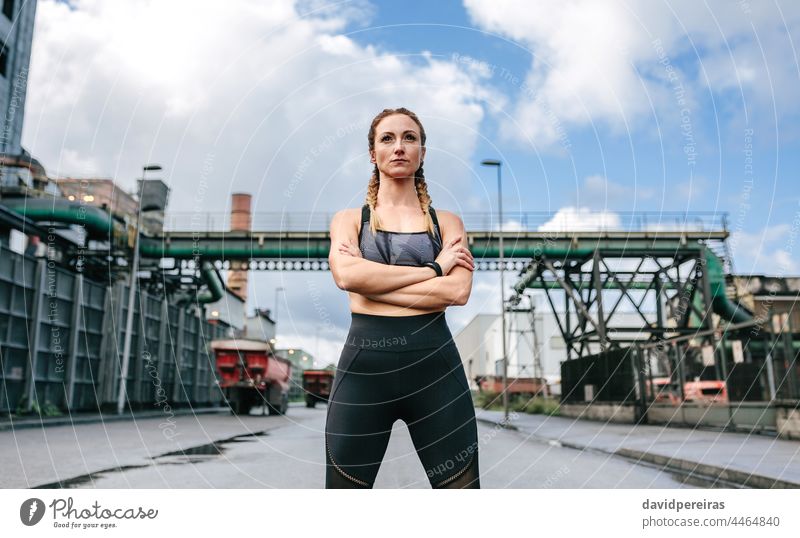 Sportliche Frau mit verschränkten Armen posiert vor einer Fabrik sportlich verschränkte Arme selbstbewußt geradeaus schauen stolz Läufer Wolken befähigt