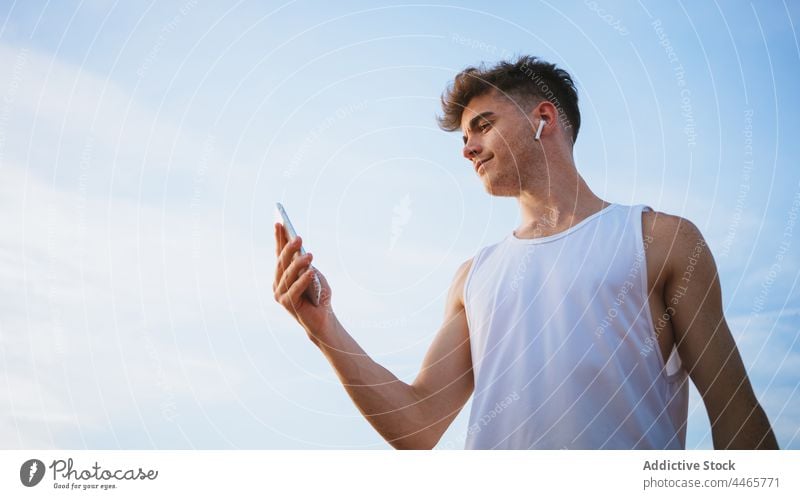 Sportler mit Smartphone, der ein Lied über einen Ohrhörer hört Ohrstöpsel zuhören Musik Gesang Wiedergabeliste genießen Himmel benutzend Apparatur Mann Gerät