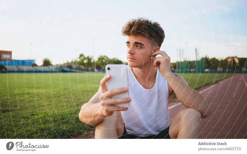 Sportler, der auf der Rennstrecke ein Lied aus einem Ohrhörer hört Athlet Ohrstöpsel zuhören Musik Gesang Smartphone Telefon Mobile nachdenken Leichtathletik