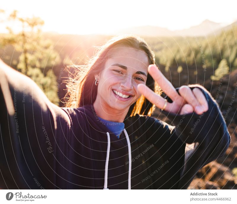 Lächelnde Teenager zeigen Frieden Geste während der Aufnahme von Selfie gegen Berg zwei Finger Selbstportrait Sieg v-Zeichen cool Berge u. Gebirge freundlich
