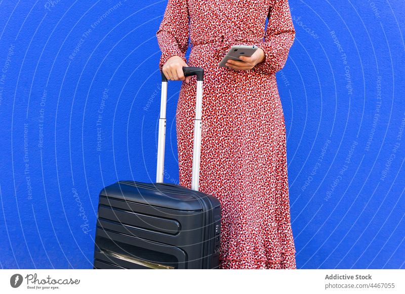Anonyme Frau steht mit Koffer und surft auf einem Mobiltelefon vor einer blauen Wand Straße Browsen Gepäck Textnachricht Smartphone positiv Großstadt rot stehen
