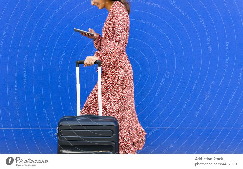 Anonyme Frau geht mit Koffer und surft auf dem Handy gegen eine blaue Wand Spaziergang Straße Browsen Gepäck Textnachricht Smartphone positiv Großstadt rot