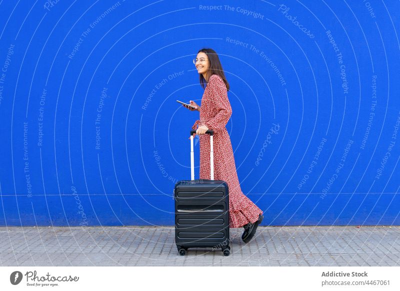 Fröhliche Frau geht mit Koffer und Handy gegen eine blaue Wand Spaziergang Straße Smartphone Gepäck Browsen positiv Großstadt rot trendy Talkrunde Apparatur